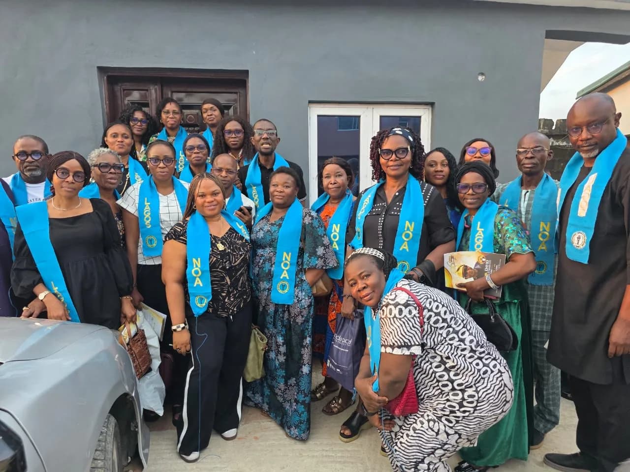 Members wearing blue NOA Lagos sashes, standing together outside a building.